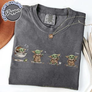 K&ouml;nnte beinhalten: Ein graues Comfort Colors T-Shirt mit einer Grafik eines gr&uuml;nen Alien-Charakters in verschiedenen Posen. Das Design zeigt den Alien in einer Kapsel, der mit einem Ball spielt, verwirrt ist und "coo!" sagt. Eine silberne Halskette mit Anh&auml;nger ist ebenfalls sichtbar.