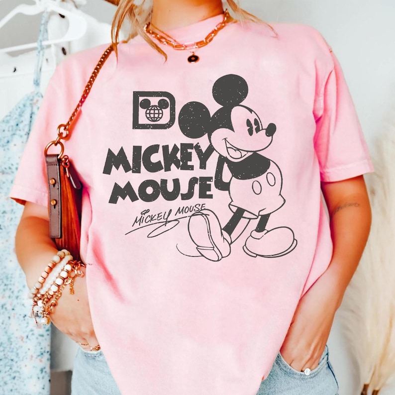 May include: Pink t-shirt featuring a black graphic of Mickey Mouse with the text "Mickey Mouse". The design includes a vintage-style Mickey Mouse illustration and signature. The shirt is a light pink color.