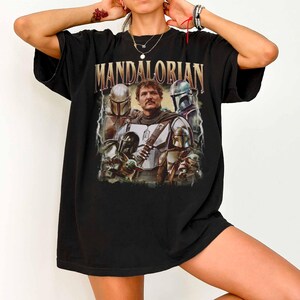 K&ouml;nnte beinhalten: Schwarzes T-Shirt mit einer Grafik des Mandalorianers und dem Wort "MANDALORIAN" in goldfarbenen Buchstaben. Die Grafik enth&auml;lt Bilder des Mandalorianers, anderer Charaktere und den Text "MANDALORIAN".