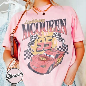May include: Pink t-shirt featuring a graphic of Lightning McQueen from the movie Cars. The design includes the text "Lightning McQueen 95" with a checkered flag and a cartoon image of the race car.
