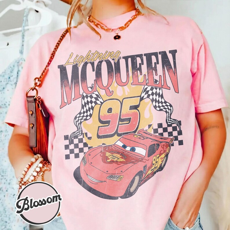 May include: Pink t-shirt featuring a graphic of Lightning McQueen from the movie Cars. The design includes the text "Lightning McQueen 95" with a checkered flag and a cartoon image of the race car.