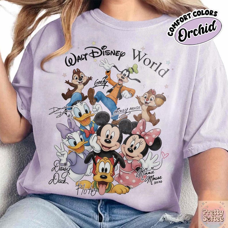 May include: A light purple t-shirt featuring a graphic of Disney characters, including Mickey Mouse, Minnie Mouse, Donald Duck, and Goofy. The text "Walt Disney World" is above the characters. The shirt also has the text "COMFORT COLORS Orchid" and "Pretty Softee".
