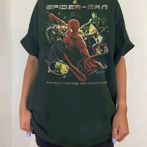 May include: Dark green Spider-Man t-shirt featuring a graphic of Spider-Man and the Green Goblin. The text "SPIDER-MAN" is at the top, and "WITH GREAT POWER COMES GREAT RESPONSIBILITY" is at the bottom. The shirt has rolled-up sleeves.