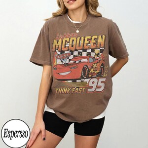 Peut inclure: T-shirt marron avec un motif de Flash McQueen du film Cars. Le graphisme comprend le texte "Lightning McQueen", "Cars", "95" et "Think Fast". Le t-shirt a un aspect vintage et vieilli.