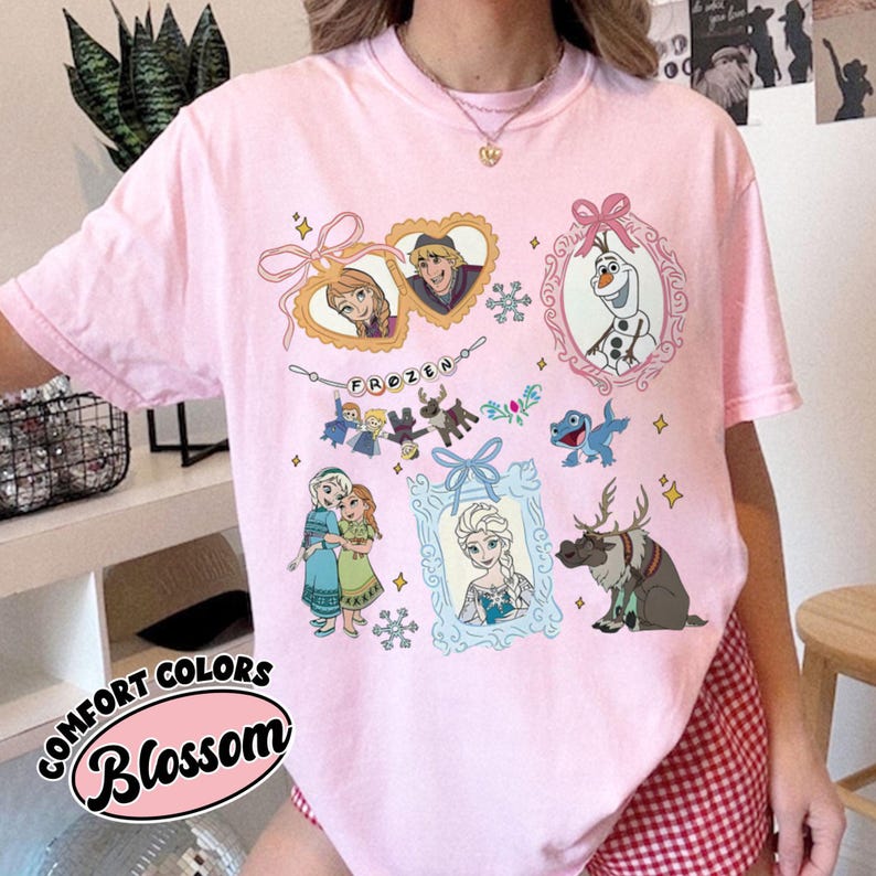 May include: A light pink t-shirt featuring various characters and elements from the movie "Frozen." The design includes framed images of Anna, Elsa, Olaf, and Kristoff, along with snowflakes and the word "FROZEN."