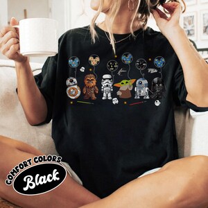 Peut inclure: T-shirt noir avec des personnages de Star Wars, dont BB-8, Chewbacca, un Stormtrooper, Grogu, R2-D2 et Dark Vador. Chaque personnage tient un ballon avec un design Star Wars. Le t-shirt porte l'inscription "Comfort Colors Black" dans un cercle.