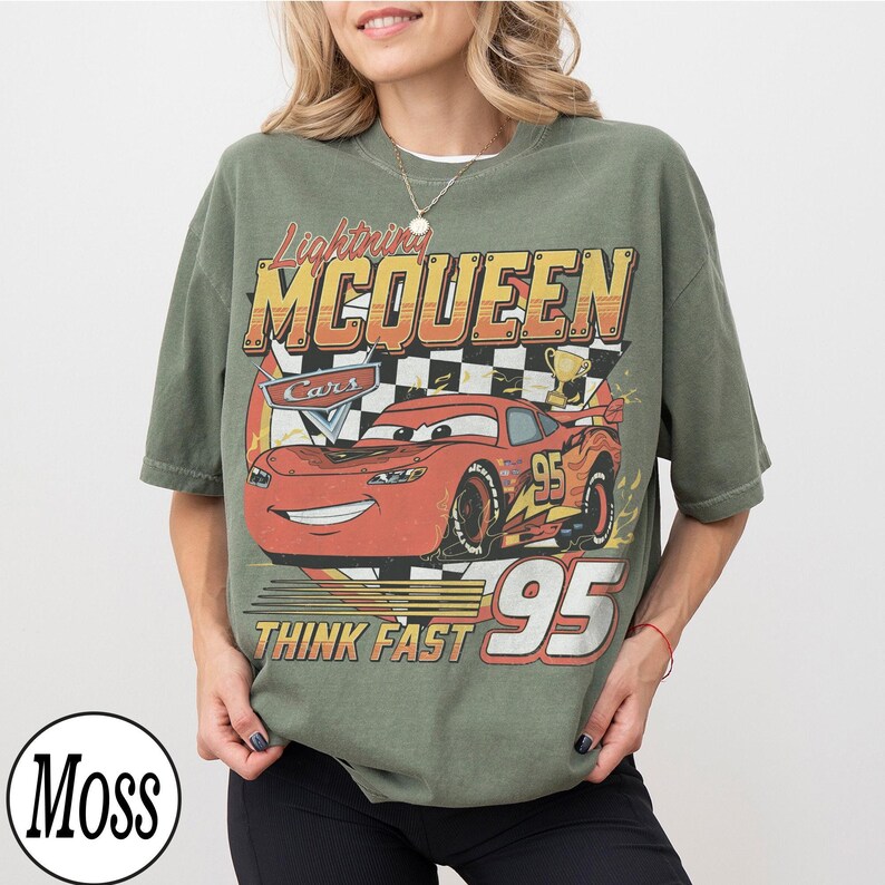 Peut inclure: T-shirt vert olive avec un motif de Flash McQueen du film Cars. Le design comprend le texte "Lightning McQueen", "Cars", "Think Fast" et le num&eacute;ro "95". Le t-shirt a un aspect vintage et vieilli.