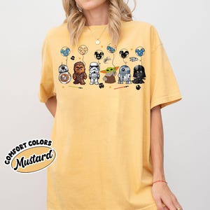Peut inclure: T-shirt moutarde avec des personnages de dessins anim&eacute;s de Star Wars et Disney. Le motif comprend BB-8, Chewbacca, un Stormtrooper, Grogu, R2-D2 et Dark Vador, chacun tenant un ballon. Le t-shirt porte les mots "Comfort Colors" et "Mustard".