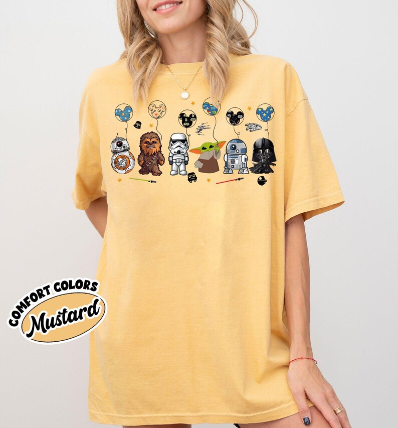 Peut inclure: T-shirt moutarde avec des personnages de dessins anim&eacute;s de Star Wars et Disney. Le motif comprend BB-8, Chewbacca, un Stormtrooper, Grogu, R2-D2 et Dark Vador, chacun tenant un ballon. Le t-shirt porte les mots "Comfort Colors" et "Mustard".
