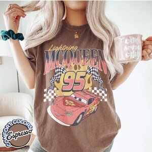 May include: Brown t-shirt featuring a graphic of Lightning McQueen from the movie Cars. The design includes the text "Lightning McQueen 95" with checkered flags and a cartoon image of the race car. The shirt also has the text "Comfort Colors Espresso".