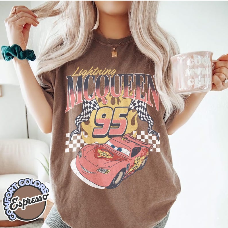 May include: Brown t-shirt featuring a graphic of Lightning McQueen from the movie Cars. The design includes the text "Lightning McQueen 95" with checkered flags and a cartoon image of the race car. The shirt also has the text "Comfort Colors Espresso".