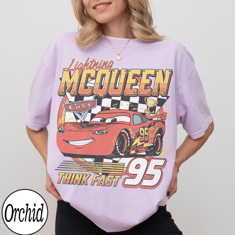 Peut inclure: T-shirt lilas avec un motif de Flash McQueen du film Cars. Le motif comprend le texte "Lightning McQueen", "Cars", "95" et "Think Fast". Le graphisme est en rouge, jaune et blanc.