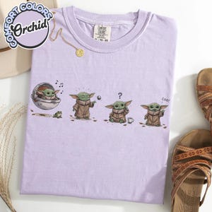 K&ouml;nnte beinhalten: Ein helllila Comfort Colors T-Shirt mit einem Cartoon eines Charakters aus einem beliebten Science-Fiction-Franchise. Der Charakter wird in vier verschiedenen Posen dargestellt, mit dem Wort "coo!" in einem Panel. Eine goldene Halskette und Sandalen sind ebenfalls sichtbar.