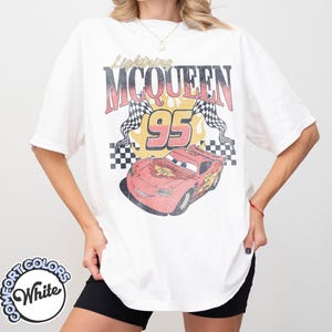 May include: White t-shirt featuring a graphic of Lightning McQueen from the movie Cars. The design includes the text "Lightning McQueen 95" with checkered flags and a red race car illustration. The shirt has short sleeves.