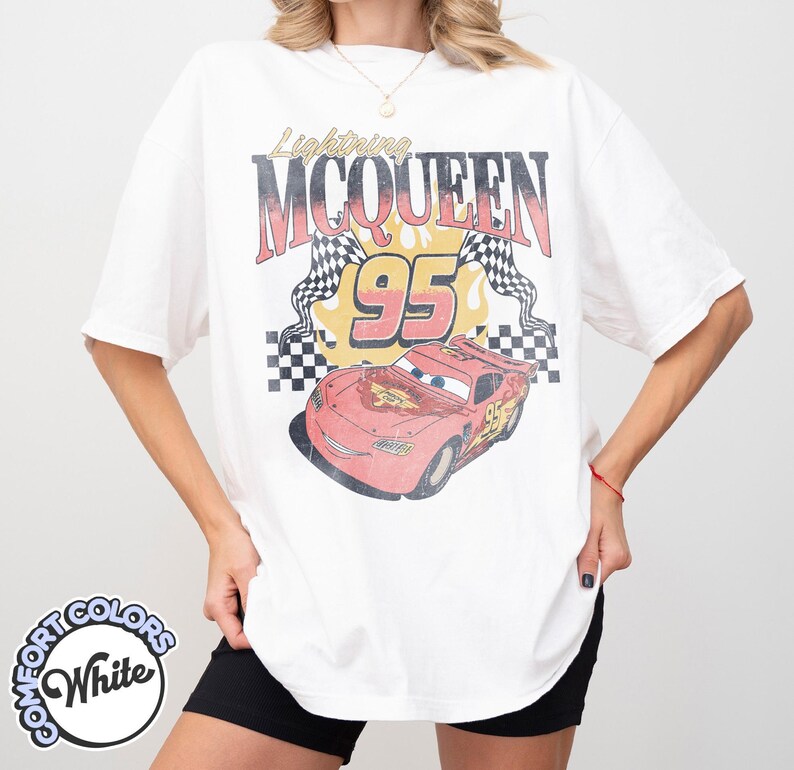 May include: White t-shirt featuring a graphic of Lightning McQueen from the movie Cars. The design includes the text "Lightning McQueen 95" with checkered flags and a red race car illustration. The shirt has short sleeves.