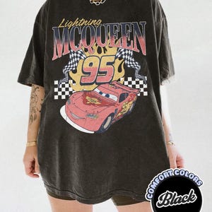 May include: Black t-shirt featuring a graphic of Lightning McQueen from the movie Cars. The design includes the text "Lightning McQueen 95" with checkered flags and flames. The shirt is a Comfort Colors brand.