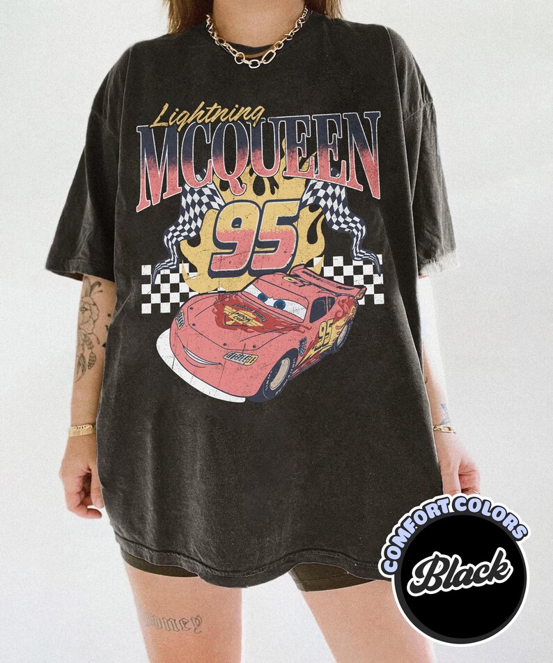 May include: Black t-shirt featuring a graphic of Lightning McQueen from the movie Cars. The design includes the text "Lightning McQueen 95" with checkered flags and flames. The shirt is a Comfort Colors brand.