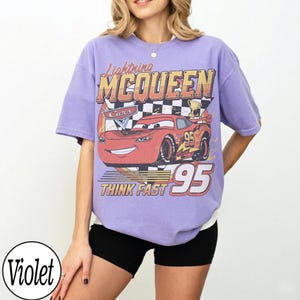 Peut inclure: T-shirt violet avec un motif de Flash McQueen du film Cars. Le design comprend le texte "Lightning McQueen", "Cars", "95" et "Think Fast". Le t-shirt a des manches courtes et une coupe ample.
