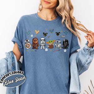 Peut inclure: T-shirt bleu jean avec des personnages de Star Wars : BB-8, Chewbacca, un Stormtrooper, Yoda, R2-D2 et Dark Vador. Le motif comprend des ballons sur le th&egrave;me des personnages et des sabres laser. Le t-shirt porte les mots "COMFORT COLORS Blue Jean" en bas &agrave; gauche.