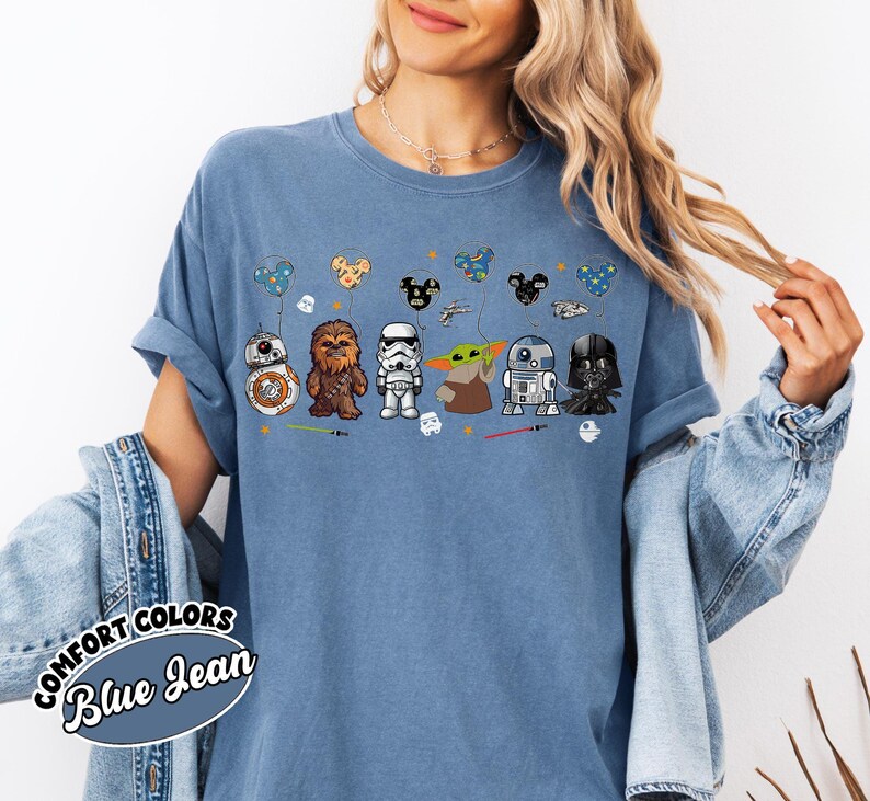 Peut inclure: T-shirt bleu jean avec des personnages de Star Wars : BB-8, Chewbacca, un Stormtrooper, Yoda, R2-D2 et Dark Vador. Le motif comprend des ballons sur le th&egrave;me des personnages et des sabres laser. Le t-shirt porte les mots "COMFORT COLORS Blue Jean" en bas &agrave; gauche.