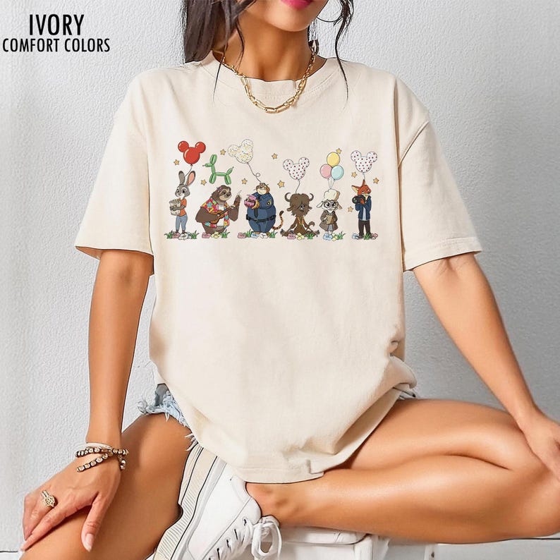 Puede incluir: Camiseta de color marfil con un gr&aacute;fico de dibujos animados de varios personajes de animales sosteniendo globos. Los globos son de varias formas y colores. El texto "IVORY COMFORT COLORS" est&aacute; impreso en la parte superior izquierda.