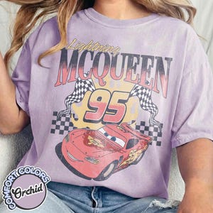 May include: Orchid-colored t-shirt featuring a graphic of Lightning McQueen from the movie Cars. The design includes the text "Lightning McQueen 95" with checkered flags and a cartoon car image.