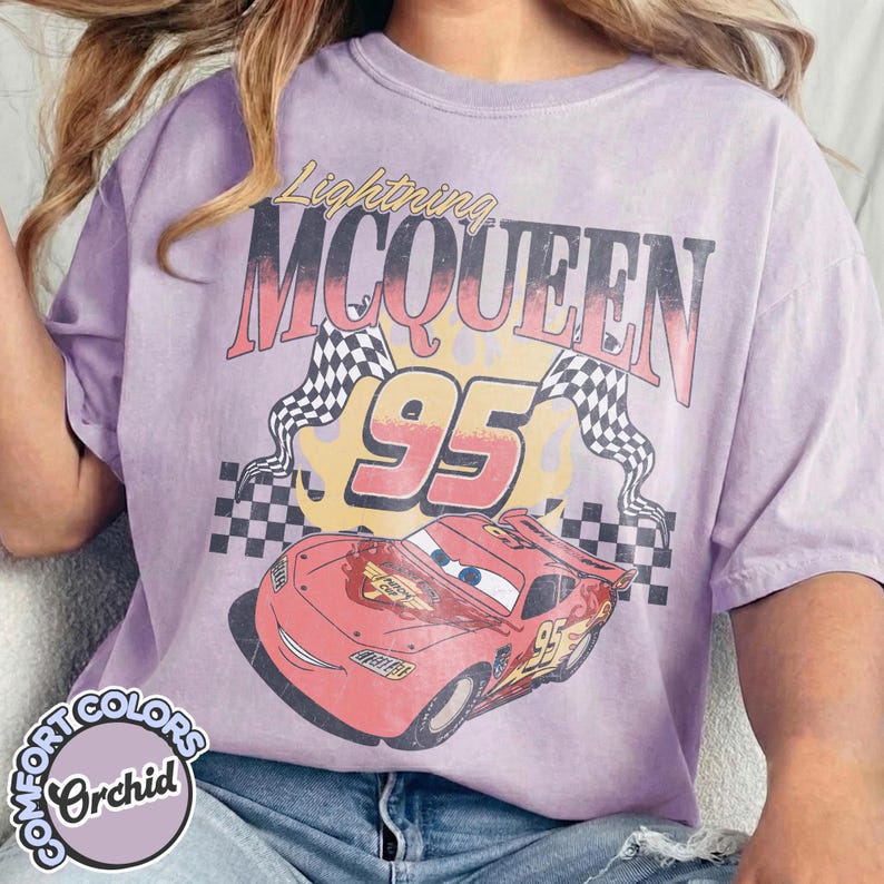 May include: Orchid-colored t-shirt featuring a graphic of Lightning McQueen from the movie Cars. The design includes the text "Lightning McQueen 95" with checkered flags and a cartoon car image.