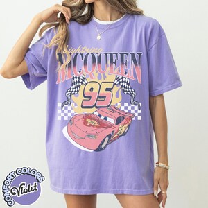 May include: A violet-colored t-shirt featuring a graphic of Lightning McQueen from the movie Cars. The design includes the text "Lightning McQueen 95" with checkered flags and a cartoon image of the race car. The shirt has a Comfort Colors label.