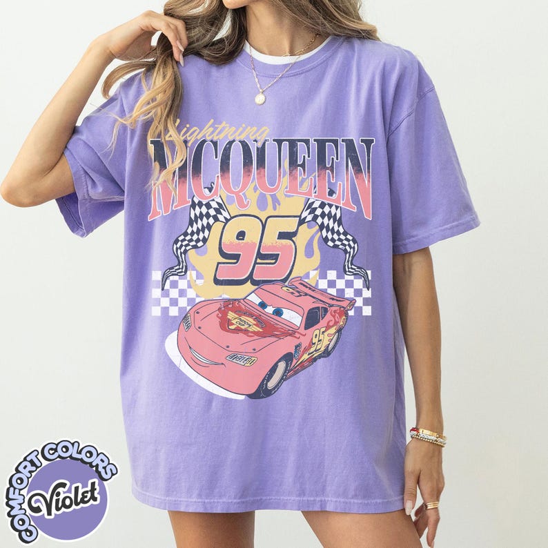 May include: A violet-colored t-shirt featuring a graphic of Lightning McQueen from the movie Cars. The design includes the text "Lightning McQueen 95" with checkered flags and a cartoon image of the race car. The shirt has a Comfort Colors label.