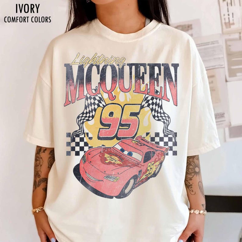 May include: Ivory-colored t-shirt featuring a graphic of Lightning McQueen from the movie Cars. The design includes the text "Lightning McQueen 95" with checkered flags and a red car illustration. The shirt is labeled "Comfort Colors".