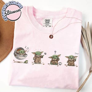 K&ouml;nnte beinhalten: Hellrosa Comfort Colors T-Shirt mit einer Grafik eines gr&uuml;nen Alien-Charakters in verschiedenen Posen. Der Charakter wird in einer Kapsel gezeigt, spielt mit einem Ball, mit einem Fragezeichen und sagt "coo!". Eine silberne Halskette und ein Logo sind ebenfalls sichtbar.