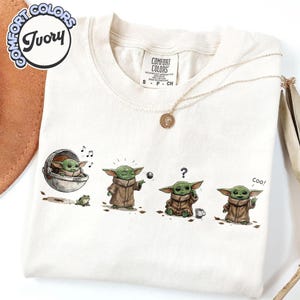 K&ouml;nnte beinhalten: Elfenbeinfarbenes T-Shirt mit einer Grafik einer Figur aus einer beliebten Science-Fiction-Serie. Die Grafik zeigt die Figur in verschiedenen Posen, mit einem Frosch, einem Ball und einer Tasse. Das Shirt hat ein Comfort Colors-Etikett.