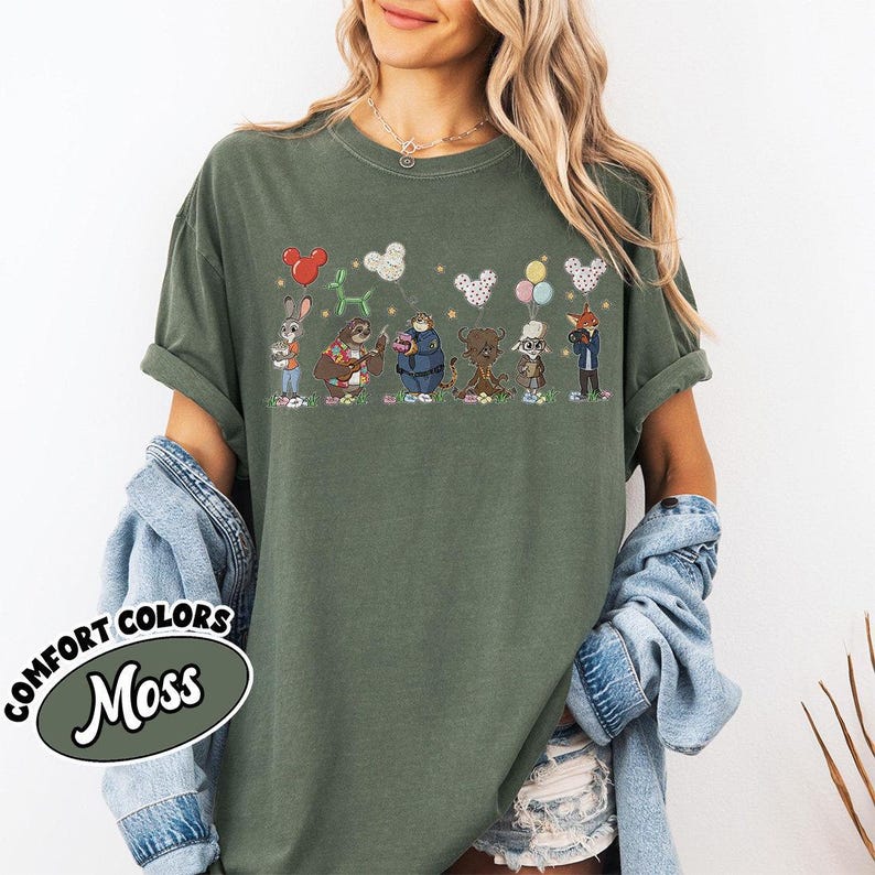 Puede incluir: Camiseta Comfort Colors verde musgo con un gr&aacute;fico caprichoso de animales de dibujos animados sosteniendo globos. El dise&ntilde;o incluye un conejo, un perezoso, un oso y un zorro. La camiseta tiene mangas enrolladas.