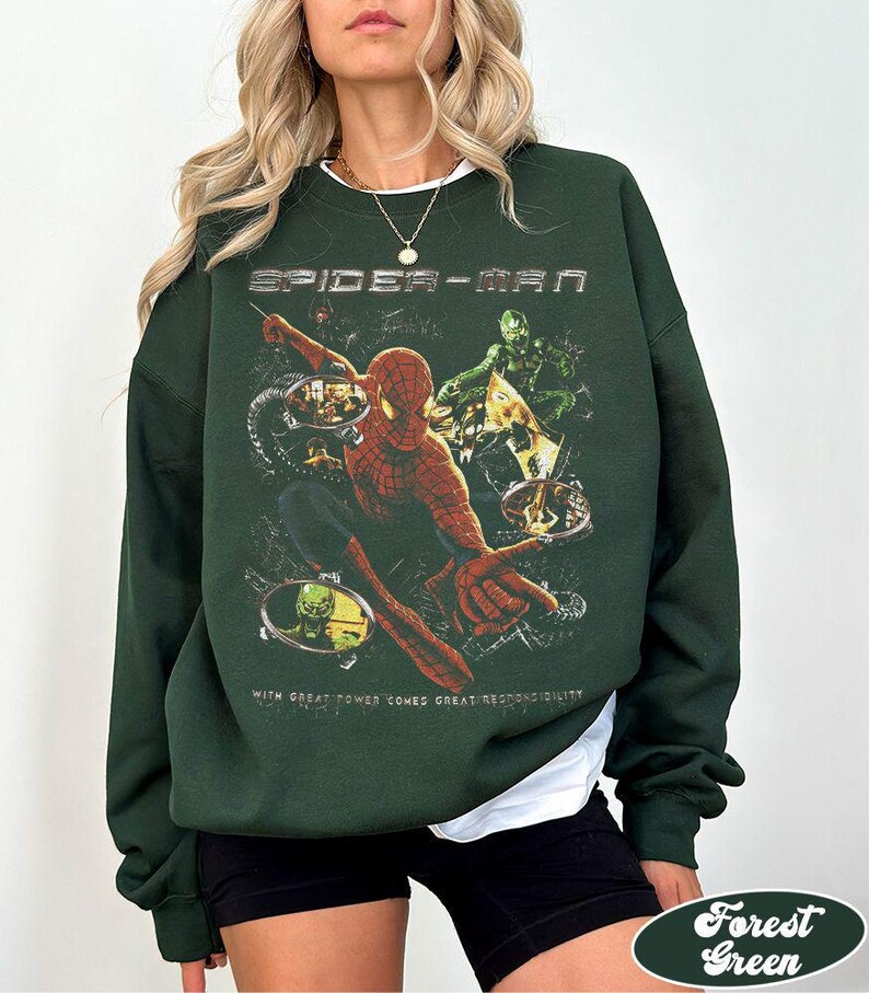 May include: Forest green sweatshirt featuring a Spider-Man graphic. The design includes the text "SPIDER-MAN" at the top and the phrase "WITH GREAT POWER COMES GREAT RESPONSIBILITY" at the bottom. The graphic depicts Spider-Man and other characters.