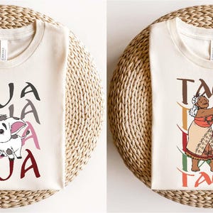 May include: Two cream-colored t-shirts with graphic designs. One shirt features a cartoon pig with the word "PUA" in gray, pink, and maroon. The other shirt displays a woman with the word "TALA" in brown, green, and maroon.