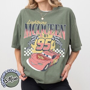 May include: Olive green t-shirt featuring a graphic of Lightning McQueen, the number 95, and checkered flags. The design includes the text "Lightning McQueen" in a vintage style. The shirt is a Comfort Colors brand.