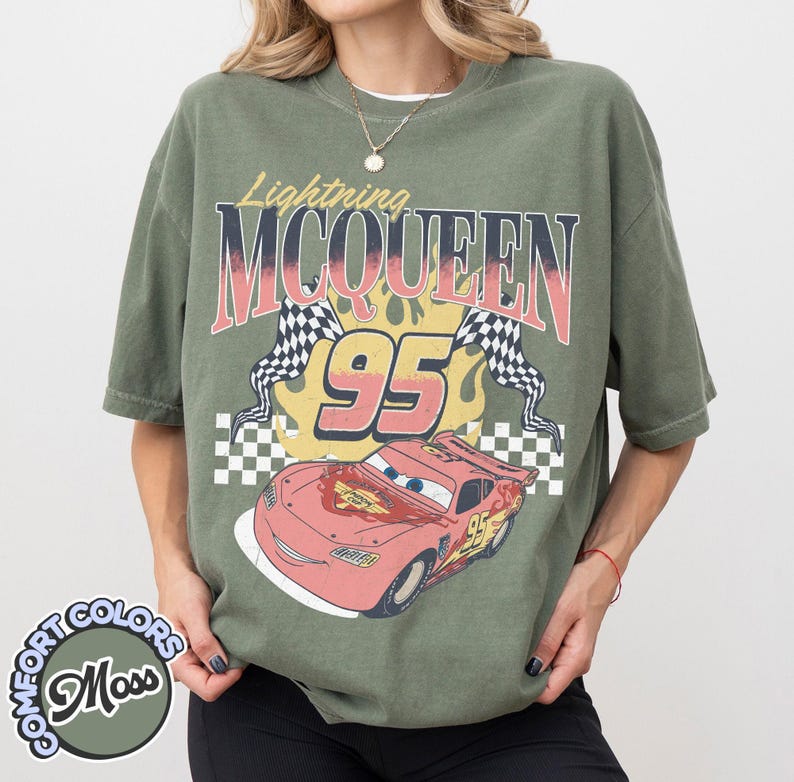 May include: Olive green t-shirt featuring a graphic of Lightning McQueen, the number 95, and checkered flags. The design includes the text "Lightning McQueen" in a vintage style. The shirt is a Comfort Colors brand.