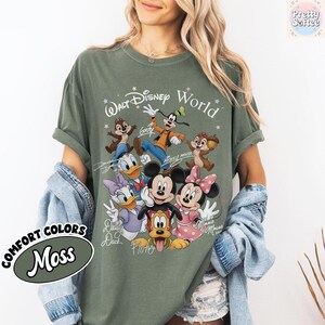 May include: Moss green t-shirt featuring a graphic of Disney characters, including Mickey and Minnie Mouse, Donald Duck, and Goofy. The text "Walt Disney World" is printed above the characters. The shirt also has the text "COMFORT COLORS" and "Moss".