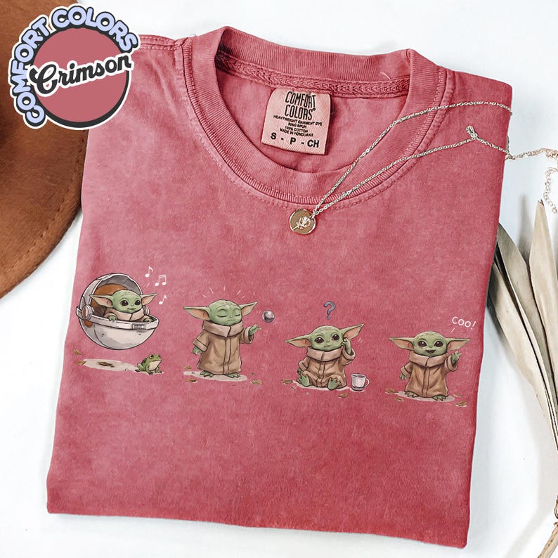 K&ouml;nnte beinhalten: Karmesinrotes T-Shirt mit einer Grafik eines Charakters in verschiedenen Posen. Der Charakter wird in einer Kapsel dargestellt, die einen Ball h&auml;lt, mit einem Fragezeichen und einem Daumen hoch. Das Shirt hat ein Comfort Colors-Etikett und eine silberne Halskette.