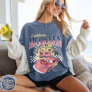 May include: A blue Lightning McQueen graphic t-shirt. The design features the text "Lightning McQueen" above the number "95" and a cartoon image of the race car. The shirt also has two checkered flags and the words "Comfort Colors Blue Jean" on the bottom left.