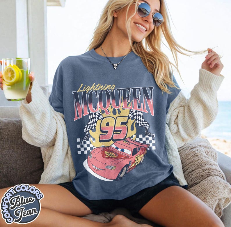 May include: A blue Lightning McQueen graphic t-shirt. The design features the text "Lightning McQueen" above the number "95" and a cartoon image of the race car. The shirt also has two checkered flags and the words "Comfort Colors Blue Jean" on the bottom left.
