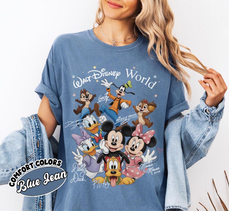 May include: Blue jean Comfort Colors t-shirt featuring a graphic of Disney characters including Mickey Mouse, Minnie Mouse, Donald Duck, and Goofy. The text "Walt Disney World" is above the characters. The shirt has rolled-up sleeves.
