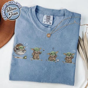 K&ouml;nnte beinhalten: Blaues T-Shirt mit einer Grafik von Grogu aus The Mandalorian. Das Design zeigt Grogu in verschiedenen Posen mit einem Frosch und einer Tasse. Auf dem Shirt stehen die Worte "Comfort Colors" und "Blue Jean". Eine goldene Halskette liegt auf dem Shirt.