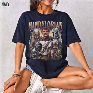 K&ouml;nnte beinhalten: Marineblaues T-Shirt mit einer Grafik von Charakteren aus der Mandalorian-Serie. Das Wort "MANDALORIAN" ist goldfarben &uuml;ber dem Bild aufgedruckt. Das T-Shirt hat einen Vintage-Look.