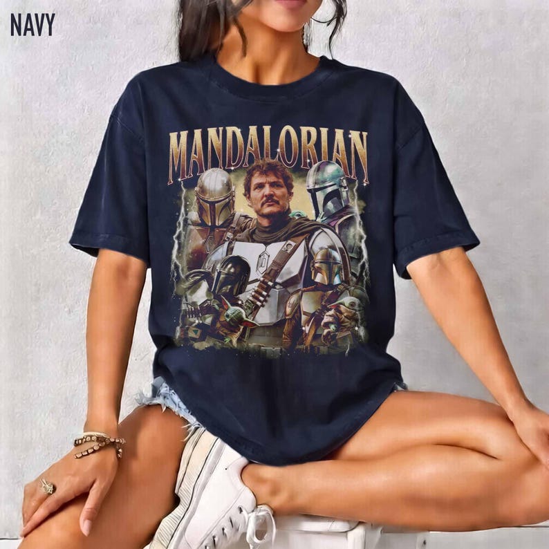 K&ouml;nnte beinhalten: Marineblaues T-Shirt mit einer Grafik von Charakteren aus der Mandalorian-Serie. Das Wort "MANDALORIAN" ist goldfarben &uuml;ber dem Bild aufgedruckt. Das T-Shirt hat einen Vintage-Look.