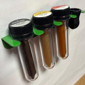 Pode incluir: Três frascos transparentes com tampas pretas, cada um contendo um pó de cor diferente, são mantidos em um suporte verde. Os frascos estão rotulados como "Truffle Noire", "Citron Confit" e "Vieux Rose".