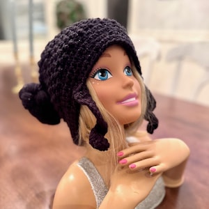 Pode incluir: Um gorro roxo escuro, feito à mão em crochê, com abas para as orelhas e um detalhe de pompom. O chapéu é exibido em uma cabeça de manequim com cabelo loiro, olhos azuis e batom rosa. O manequim está usando um top de lantejoulas prateadas.
