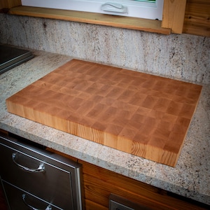 May include: A large, rectangular end-grain butcher block cutting board made of light brown wood. The board is constructed from small, square pieces of wood, creating a checkerboard pattern. It sits on a speckled countertop near a window.