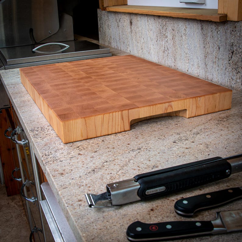 May include: A large, rectangular butcher block cutting board made of light-coloured wood. The board has a checkerboard pattern and a cutout handle. Kitchen tools, including a knife and tongs, are on the counter.