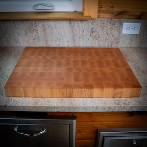 May include: A large, rectangular butcher block cutting board made of light-coloured wood. The board has a checkerboard pattern and sits on a speckled countertop. The wood grain is visible. The board appears to be new.